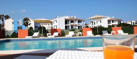 A poolside view with a glass of orange drink, white lounge chairs, and a clear blue sky.