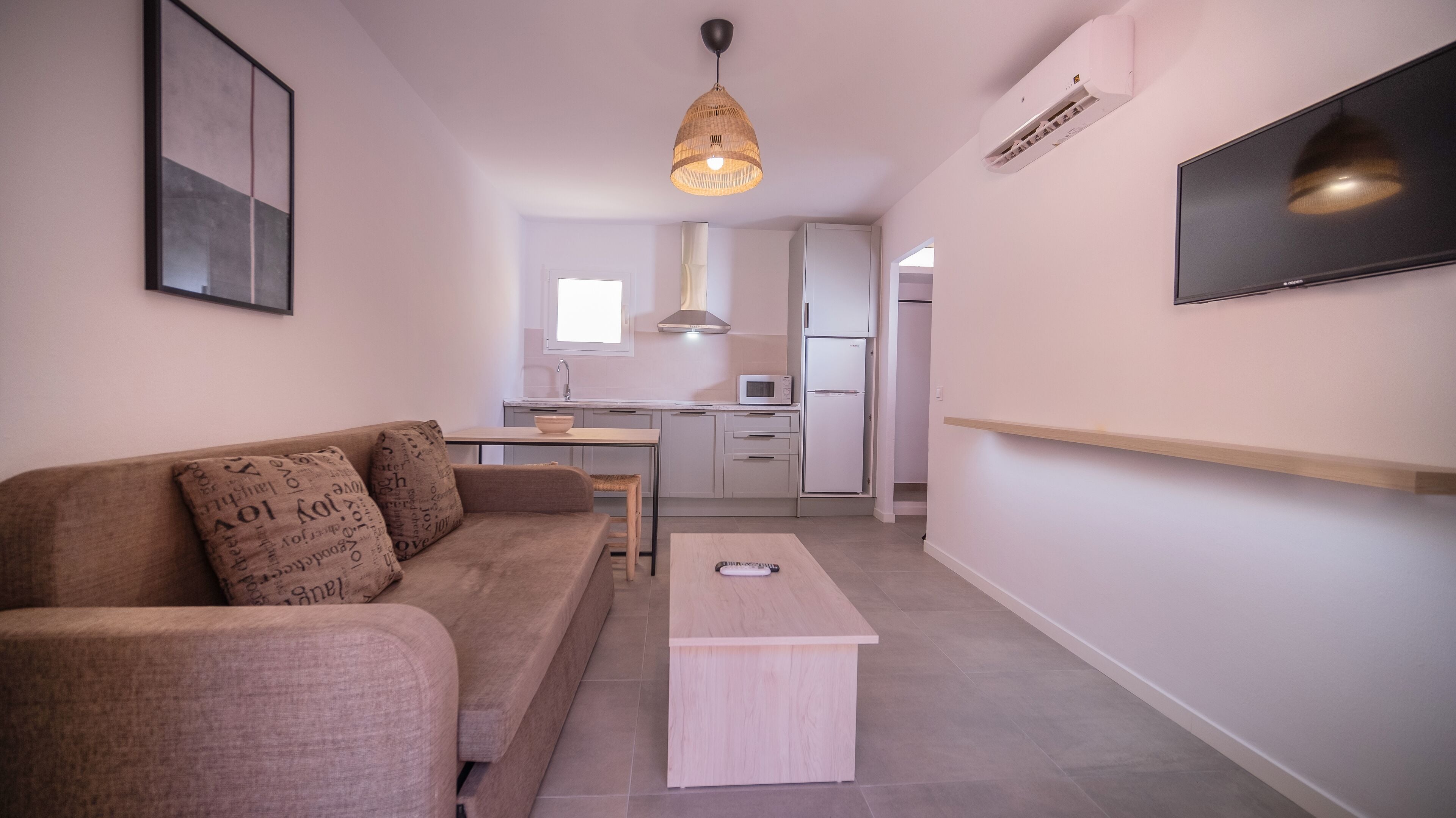 A modern living room with a sofa, a coffee table, a TV mounted on the wall, and a kitchen area in the background.