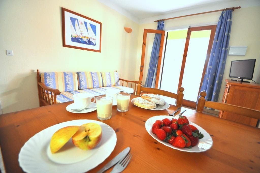 A dining area with a wooden table set for breakfast, including plates of fruit and pastries, a television, and a window with curtains.