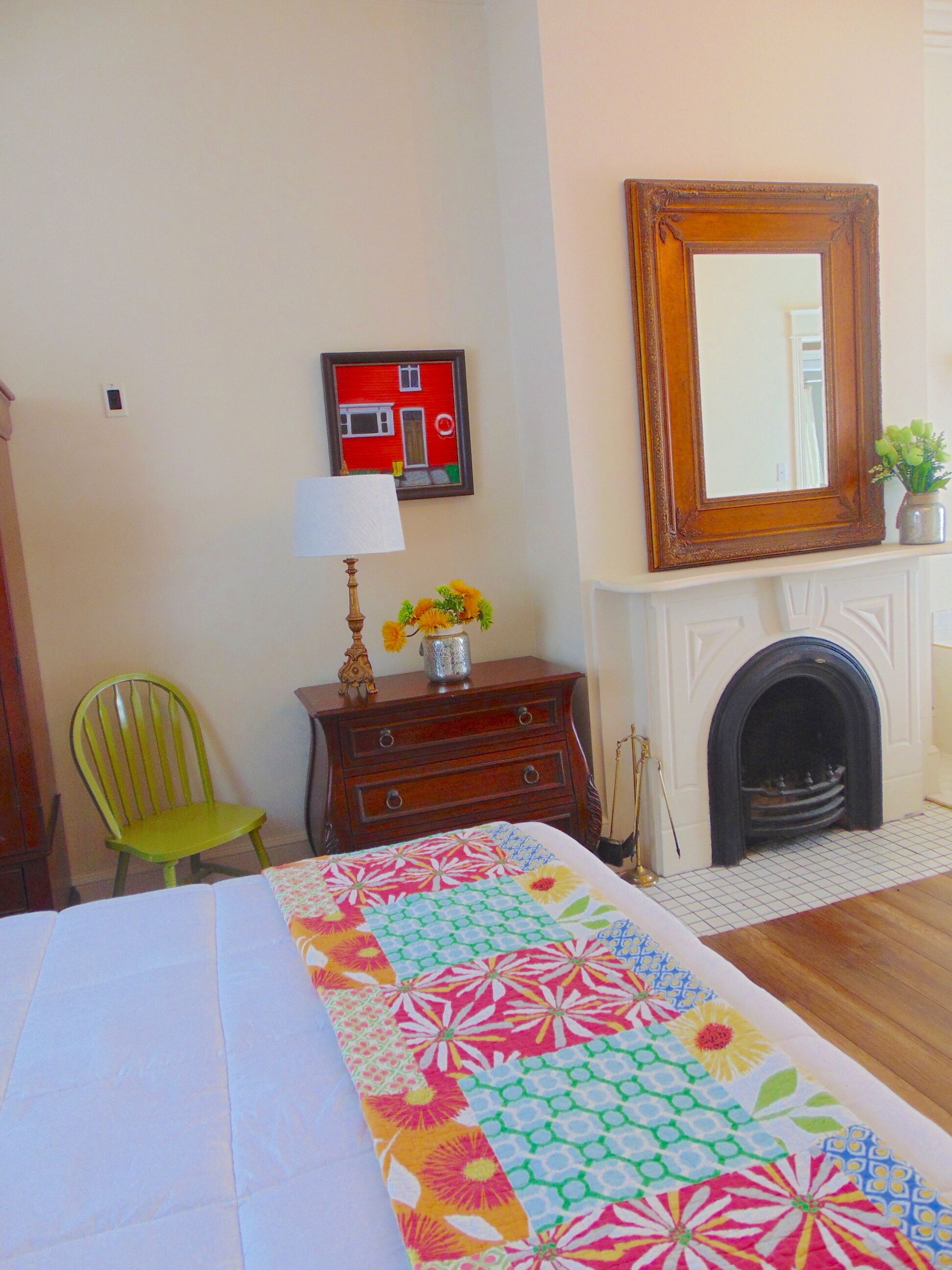 Un dormitorio con cama, cómoda, silla, chimenea y espejo montado en la pared.