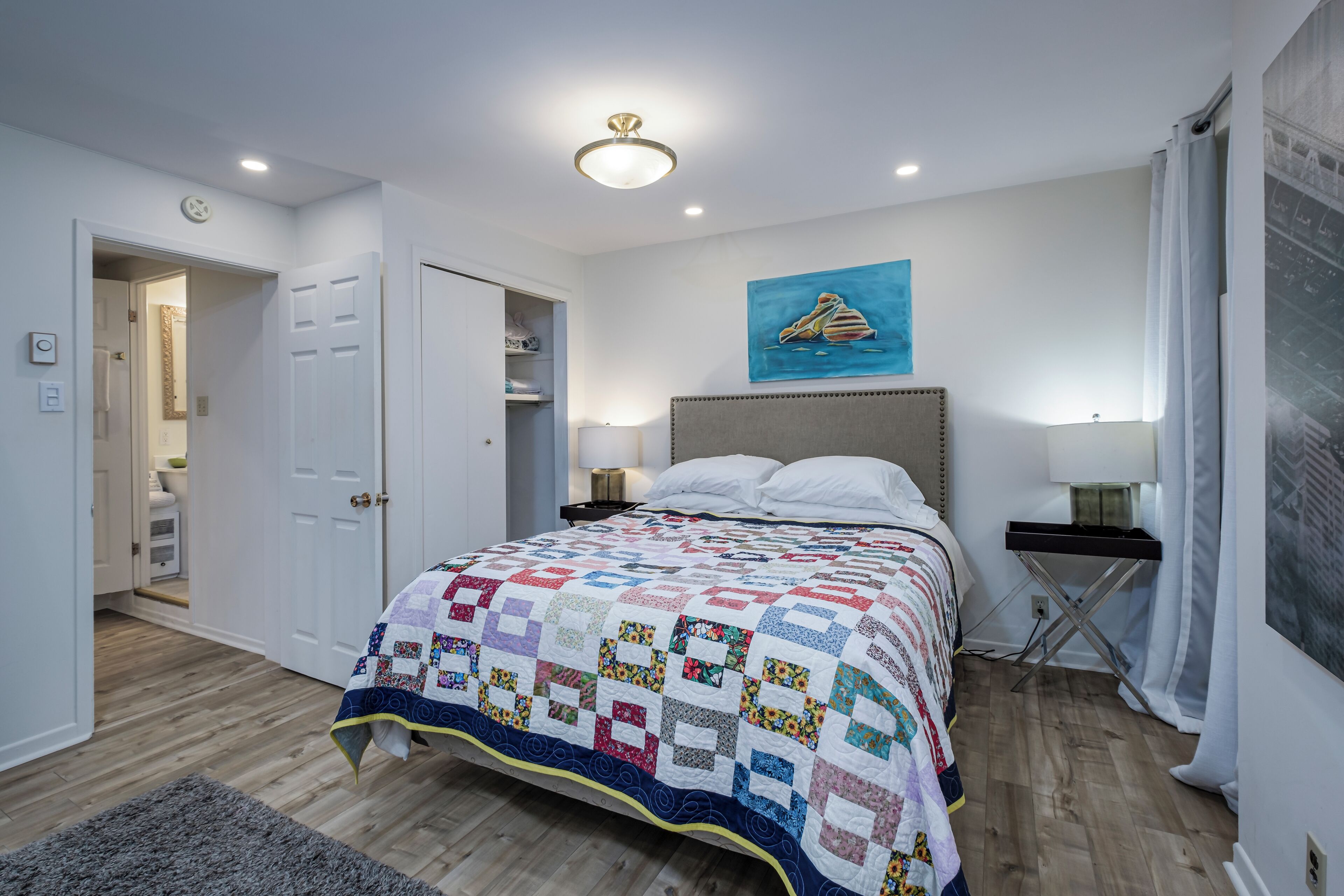 A bedroom with a large bed, a headboard, bedside lamps, a painting on the wall, and a door leading to another room.