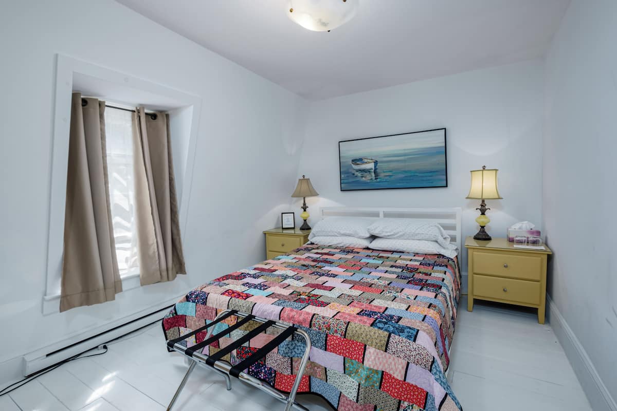 A bedroom with a bed, a nightstand, a lamp, a painting, and a window with curtains.
