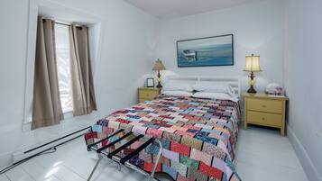 Un dormitorio con cama, mesita de noche, lámpara, cuadro y ventana con cortinas.