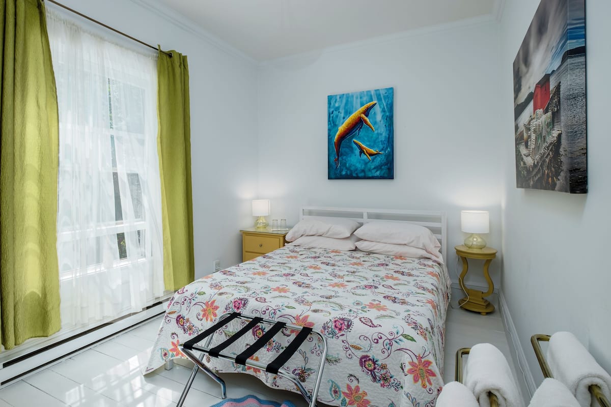 A bedroom with a bed, two bedside tables with lamps, a window with curtains, and artwork on the walls.