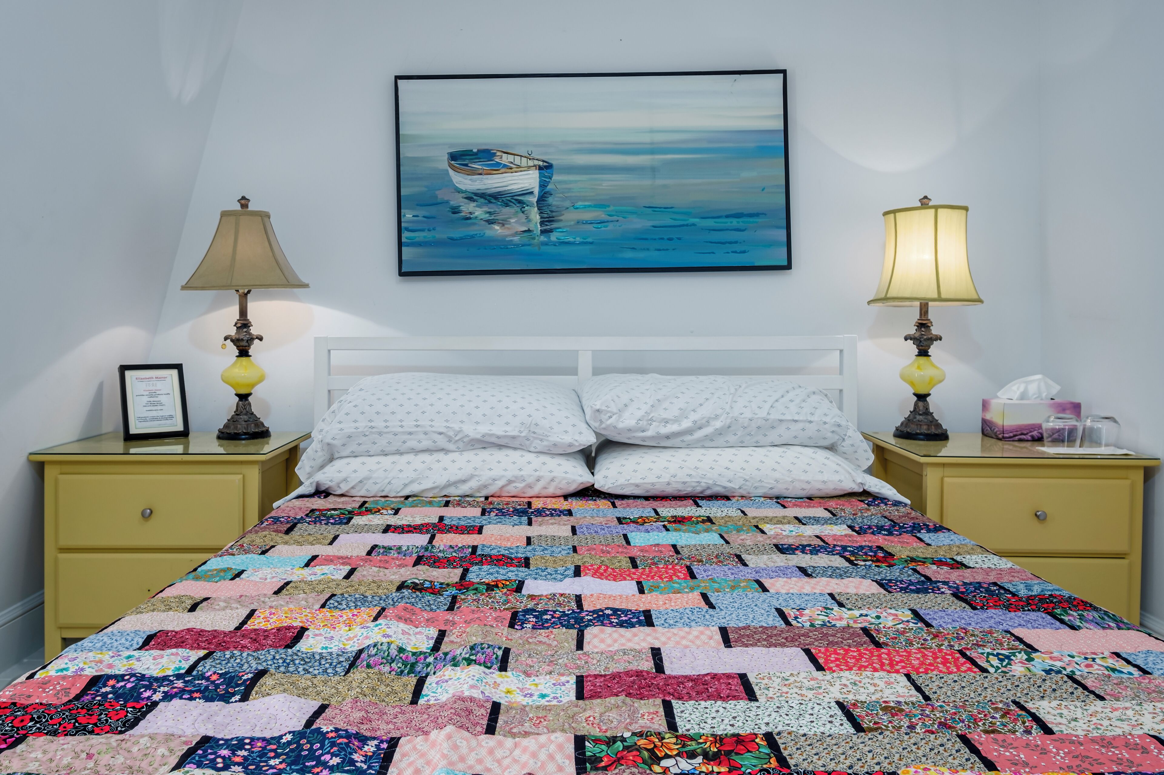 A bedroom with a colorful quilt, two bedside lamps, a framed picture, and a painting of a boat on the wall.