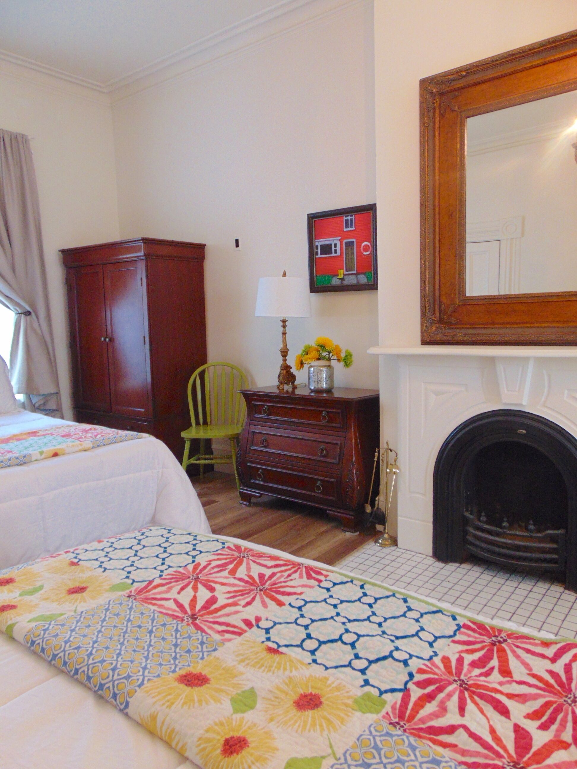 A bedroom with two beds, a dresser, a chair, a lamp, a painting, and a fireplace.