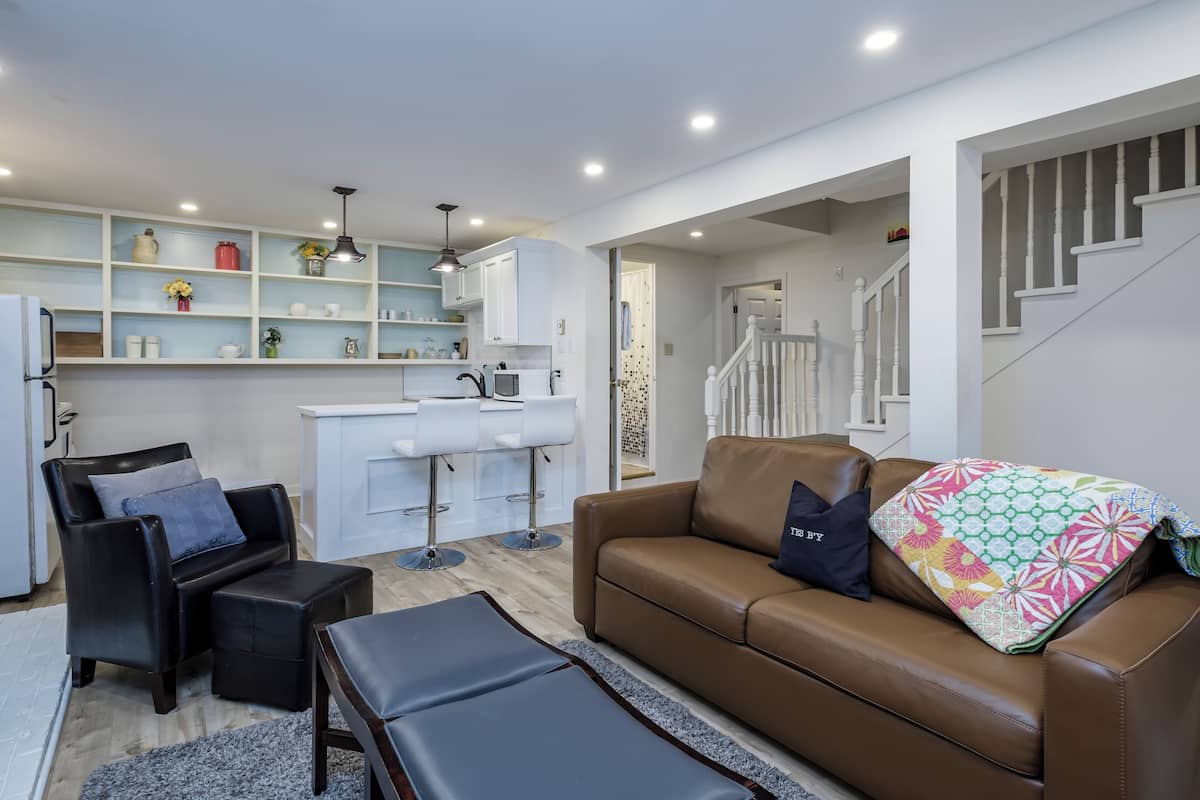 A modern living room with a brown leather sofa, a black leather armchair, and a coffee table. There is a kitchen area with white cabinets and a white countertop.