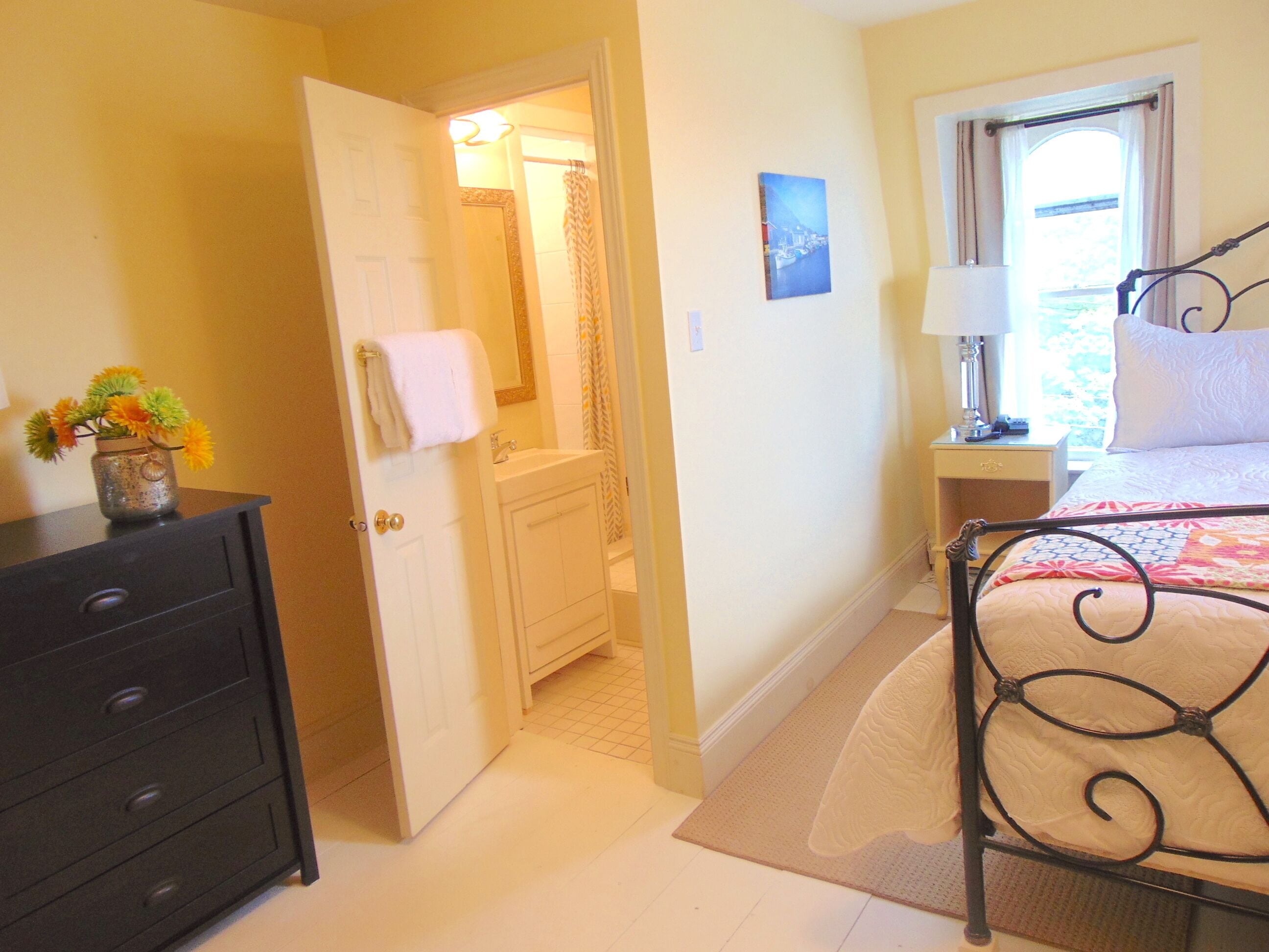 A bedroom with a bed, a nightstand, a dresser, and a bathroom visible through an open door.