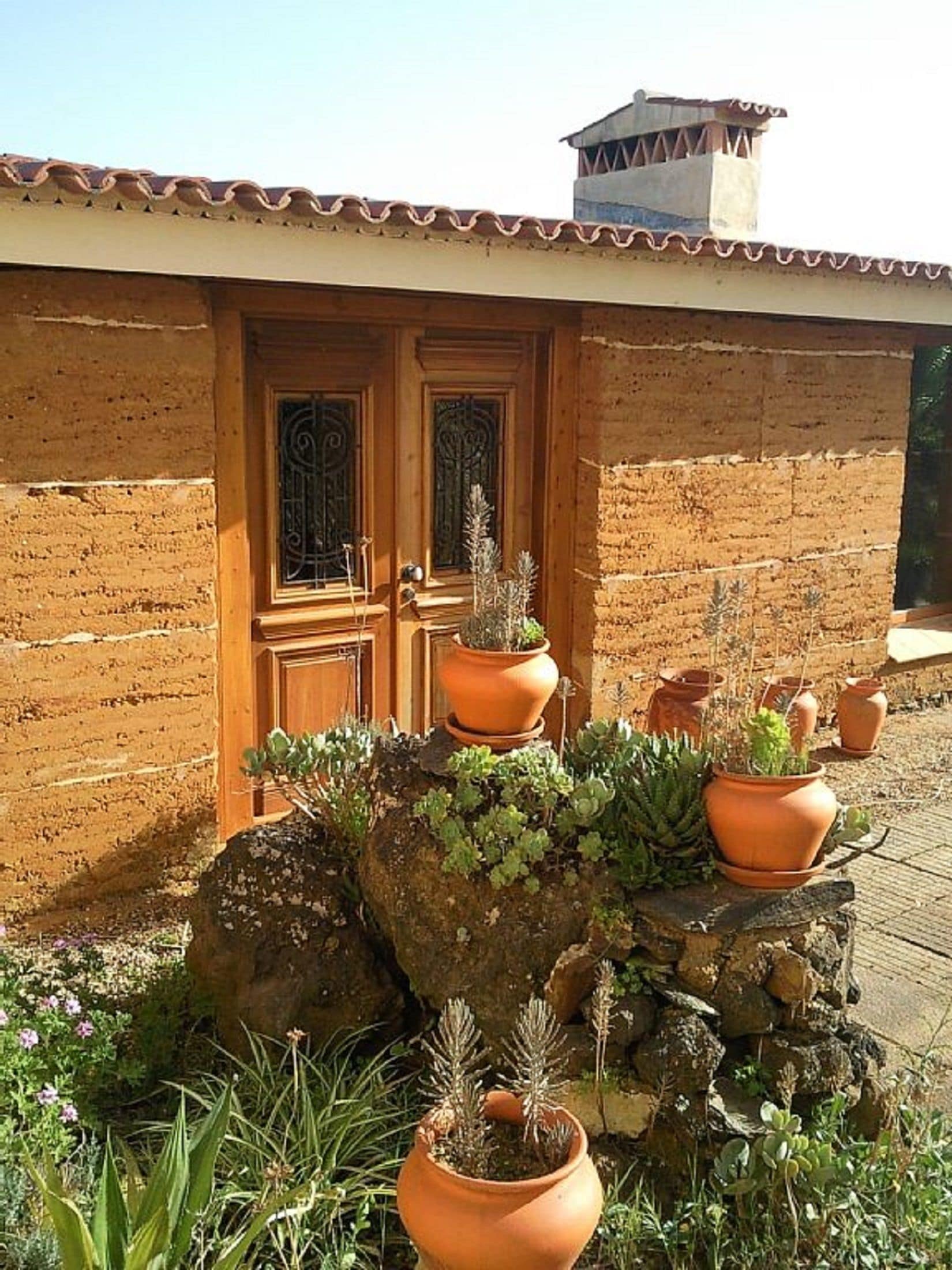A rustic building with a tiled roof and a wooden door. There are terracotta pots with plants on a rock in front of the door.