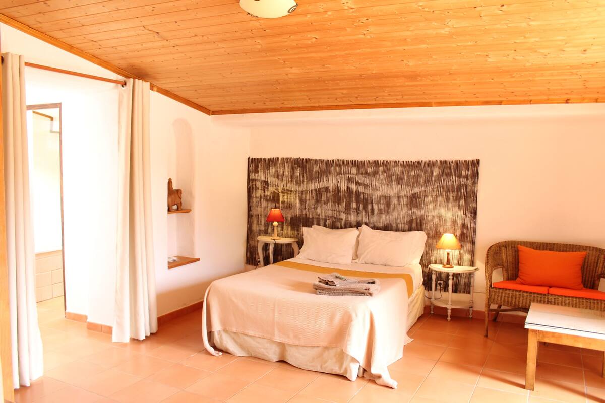 A bedroom with a wooden ceiling, a bed with a white bedspread, two bedside tables with lamps, a wicker sofa, and a small table.