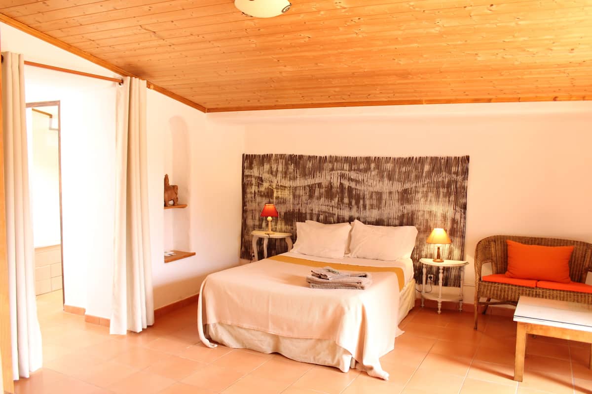 A bedroom with a wooden ceiling, a bed with a white bedspread, two bedside tables with lamps, a wicker sofa, and a small table.