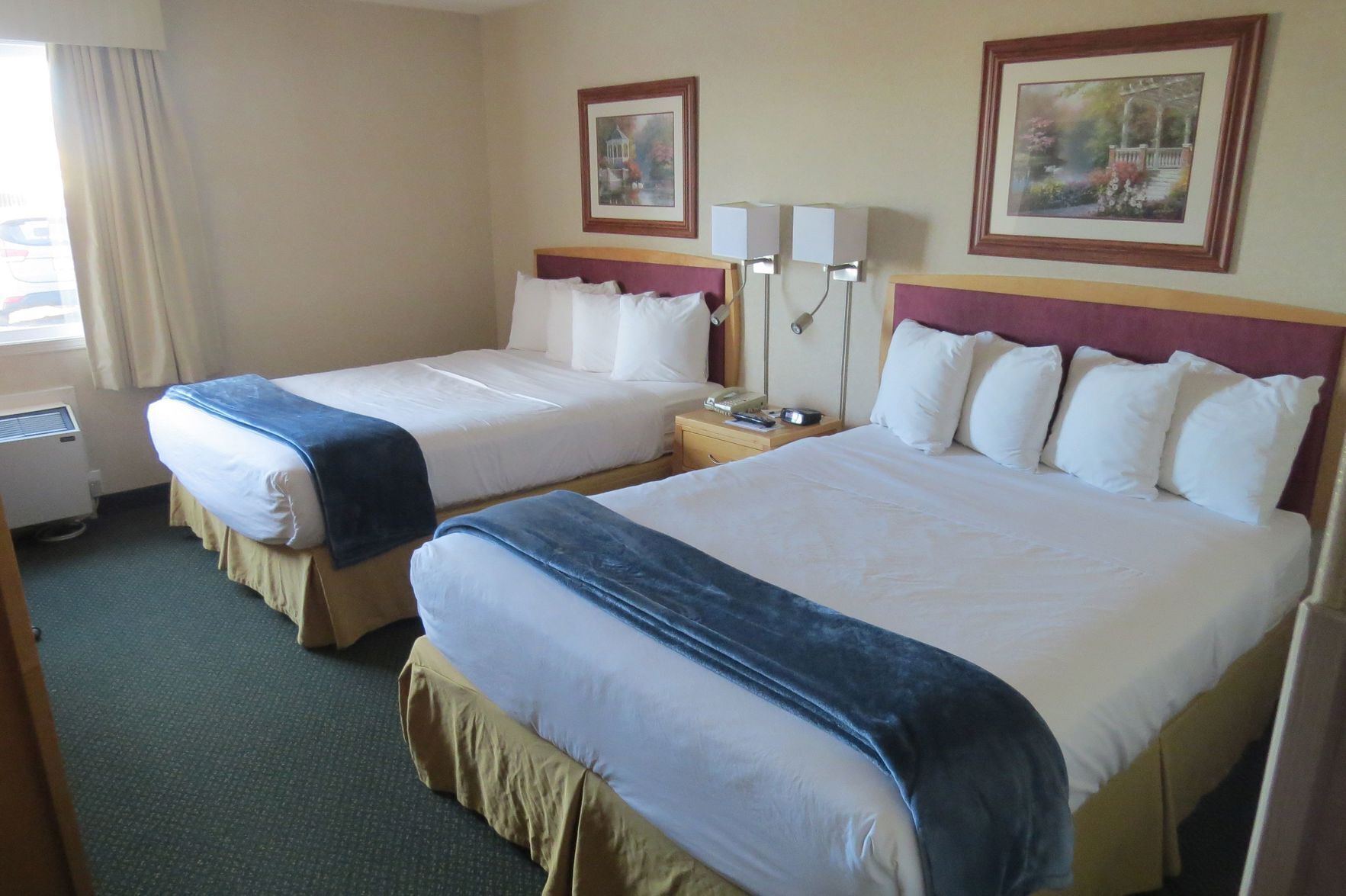A hotel room with two beds, a bedside table with a lamp, a window with curtains, and two framed pictures on the wall.