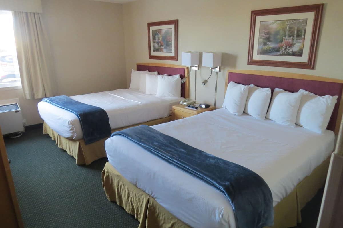 A hotel room with two beds, a bedside table with a lamp, a window with curtains, and two framed pictures on the wall.