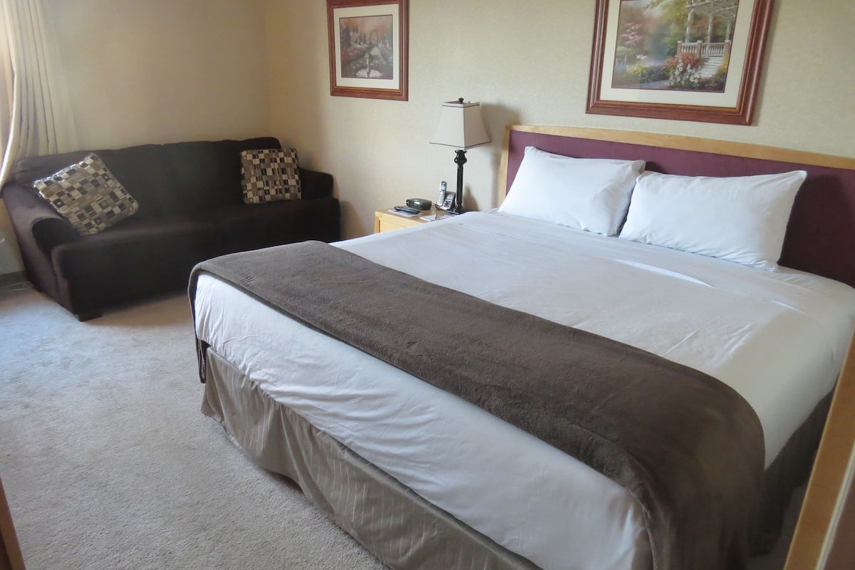 A hotel room with a large bed, a sofa, a nightstand with a lamp, and two framed pictures on the wall.