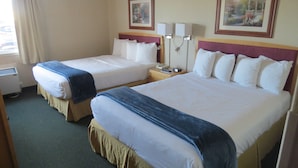 Habitación de hotel con dos camas, una mesita de noche con lámpara, un ventanal con cortinas y dos cuadros enmarcados en la pared.