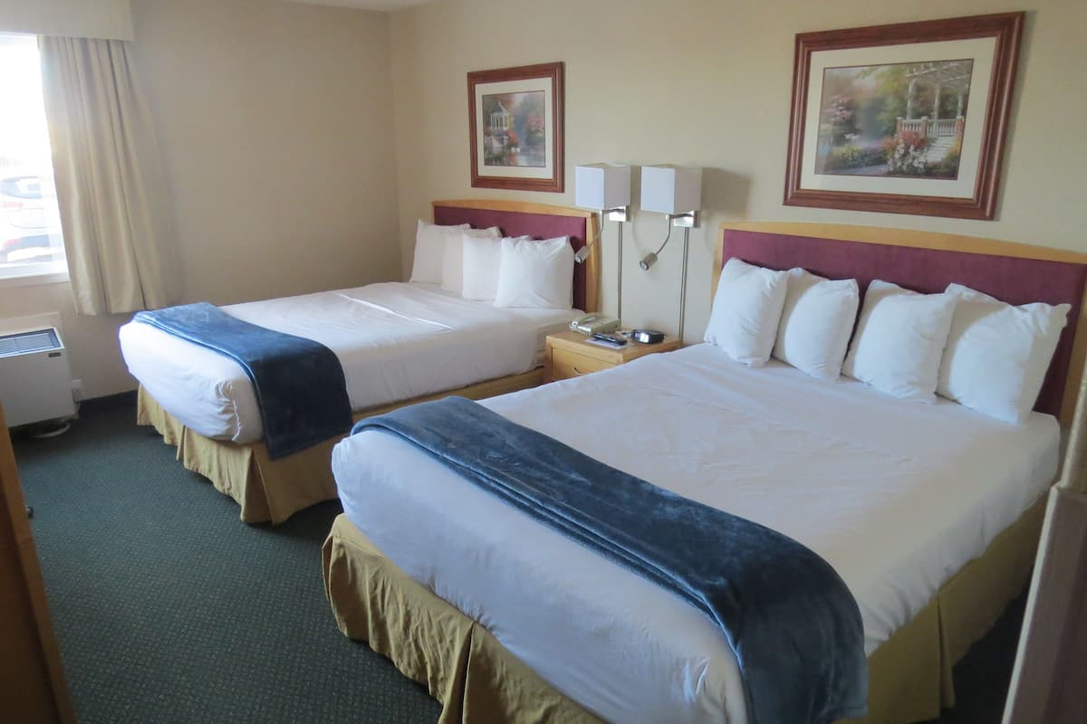 A hotel room with two beds, a bedside table with a lamp, a window with curtains, and two framed pictures on the wall.