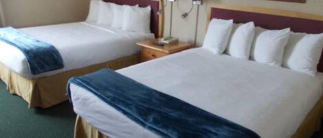 Habitación de hotel con dos camas, cada una con sábanas blancas y una manta azul. Hay dos mesitas de noche con lámparas y un teléfono. La habitación tiene alfombra verde y paredes beige.