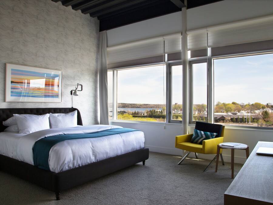 Slaapkamer met een groot bed, een gele stoel, een klein tafeltje en uitzicht op een landschap door grote ramen.