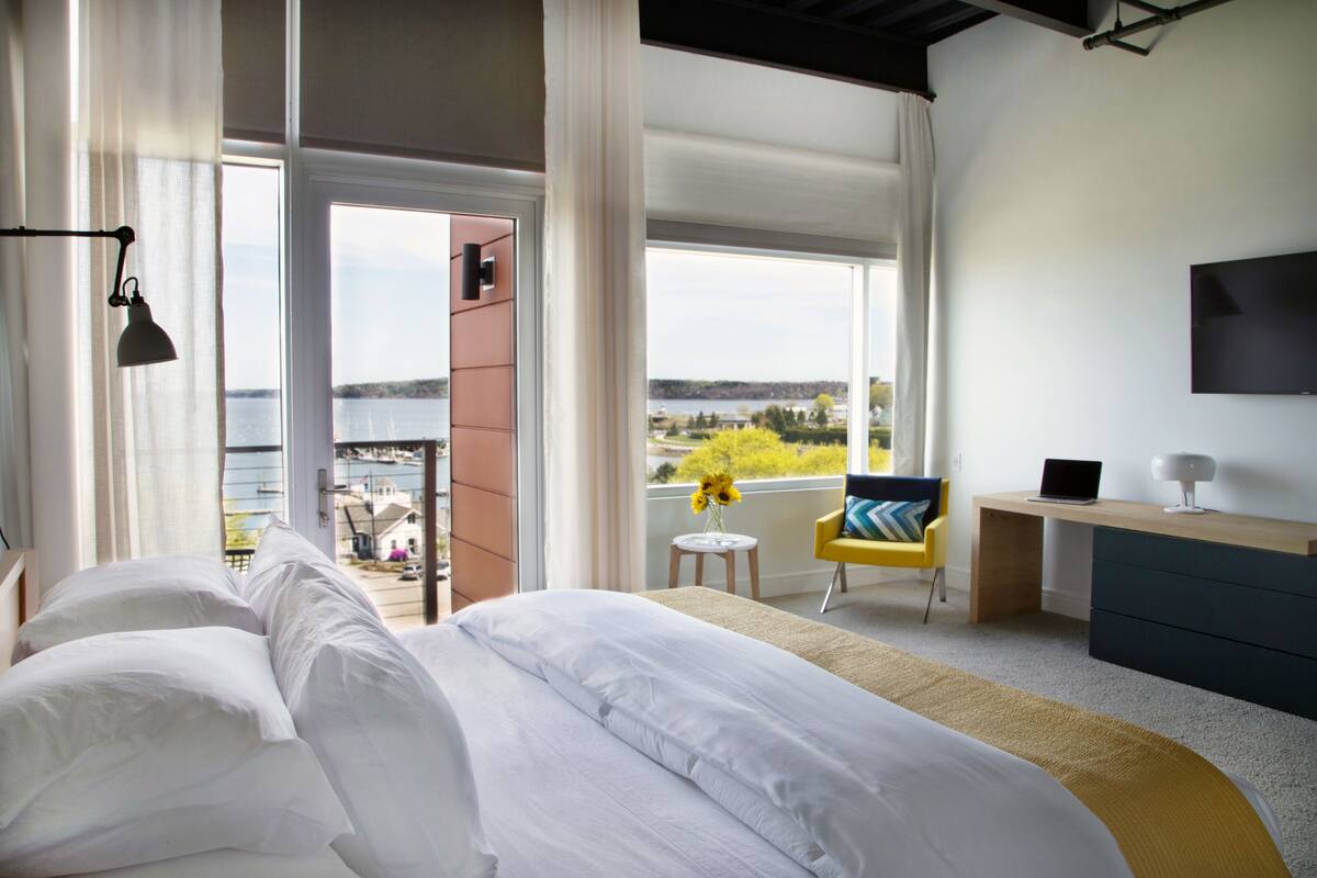 Et moderne hotelværelse med en stor seng, et skrivebord, en stol og et fjernsyn. Der er en balkon med udsigt over byen og et vandareal.