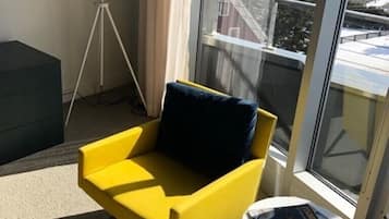 Un sillón amarillo con un cojín oscuro, una mesita blanca con revistas, y vistas a edificios a través de un ventanal grande.