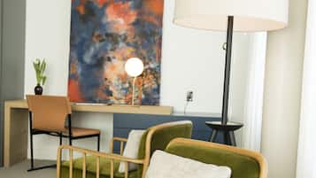 Une pièce moderne avec un fauteuil vert, une table ronde sur laquelle repose un magazine, un bureau avec une chaise, une lampe et un grand tableau abstrait accroché au mur.