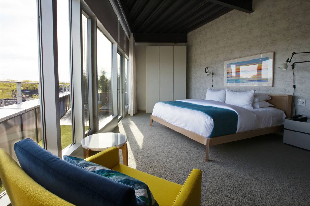 Et moderne hotelværelse med en stor seng, et lille rundt bord, en gul lænestol og en balkon med udsigt over byen.