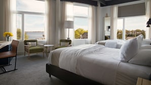 A hotel room with a large bed, two armchairs, a small table, and a desk. There are large windows with curtains, a lamp, and a view of the sea. - 250 Main Hotel (Rockland)