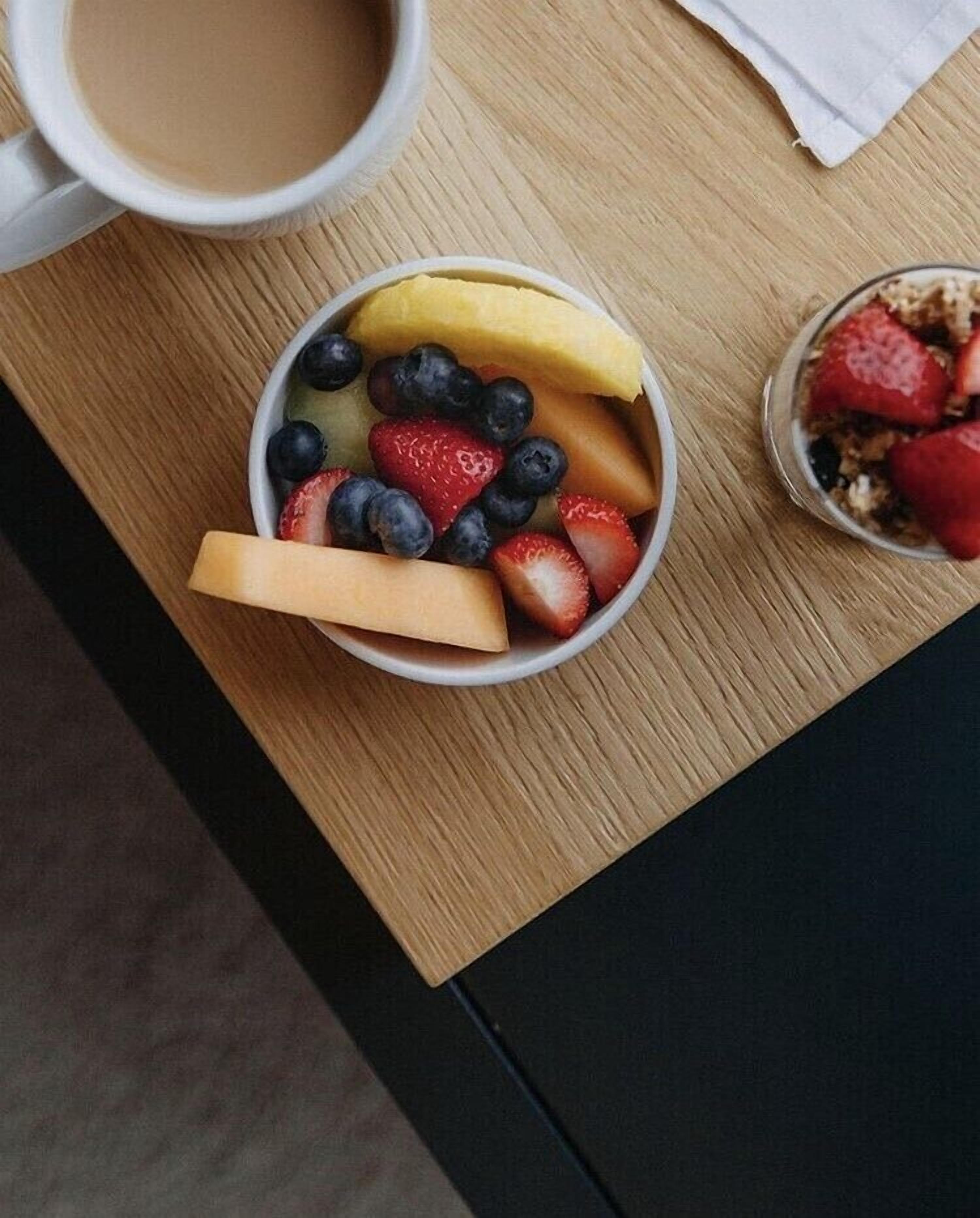 Un bol de fruta sobre una mesa de madera, con una taza de café a su lado.