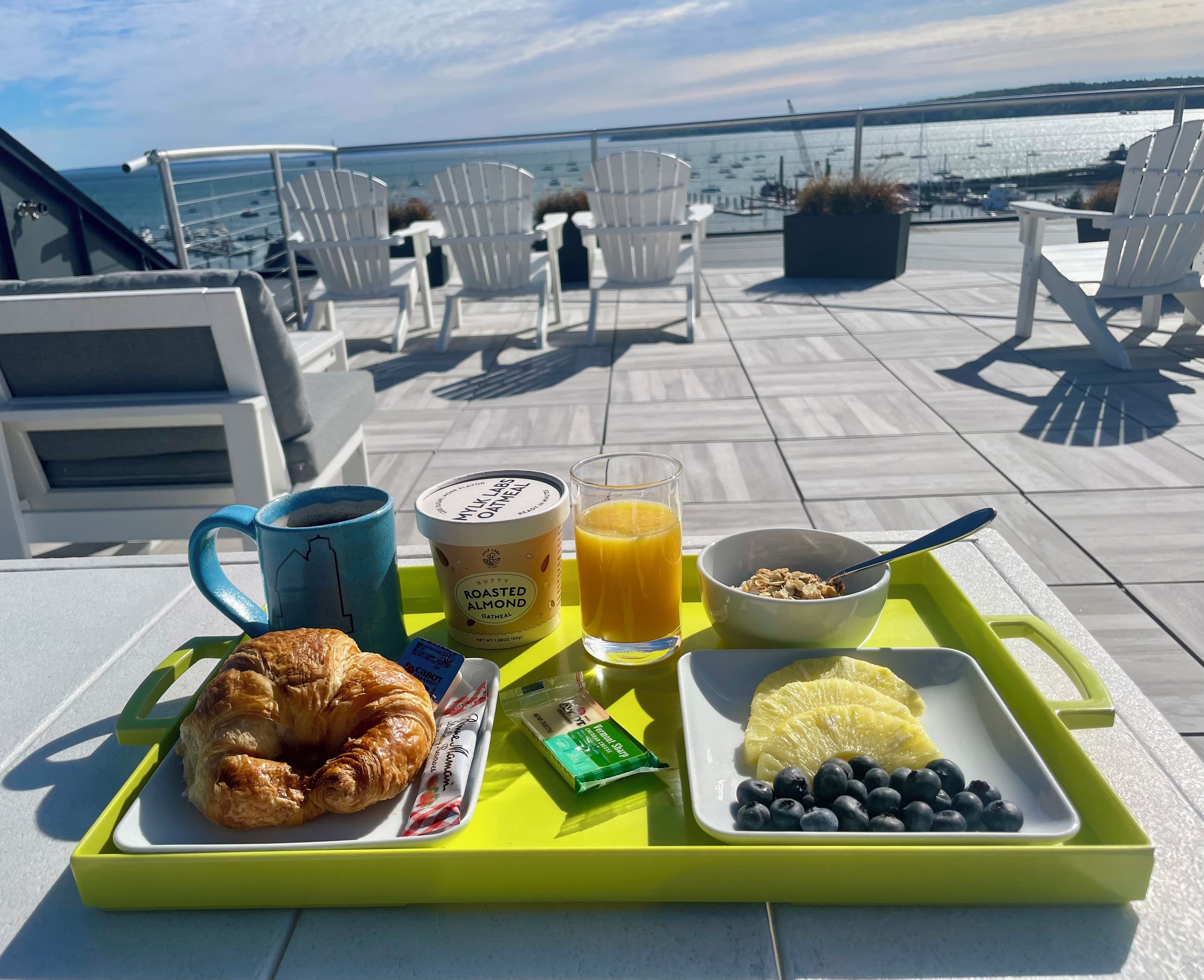 A breakfast tray with croissant, orange juice, yogurt, and blueberries on a terrace with chairs and a view of the water.