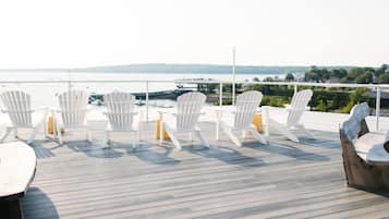 Um terraço com cadeiras Adirondack brancas e uma mesa de madeira, com vista para a água e terras distantes.