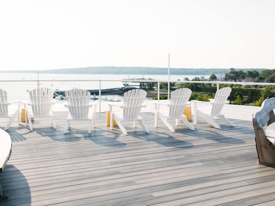 Um terraço com cadeiras Adirondack brancas e uma mesa de madeira, com vista para a água e terras distantes.
