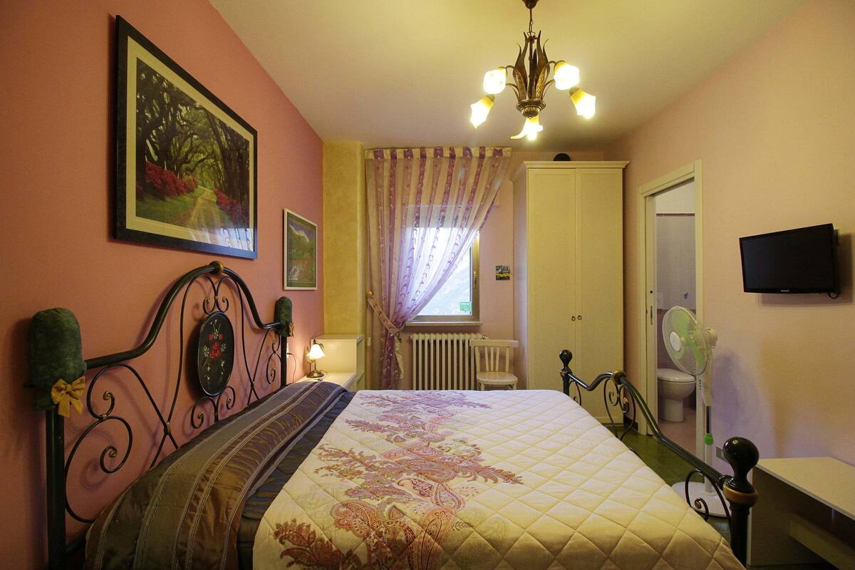 A bedroom with a bed, a radiator, a window with curtains, a fan, and a television on the wall.