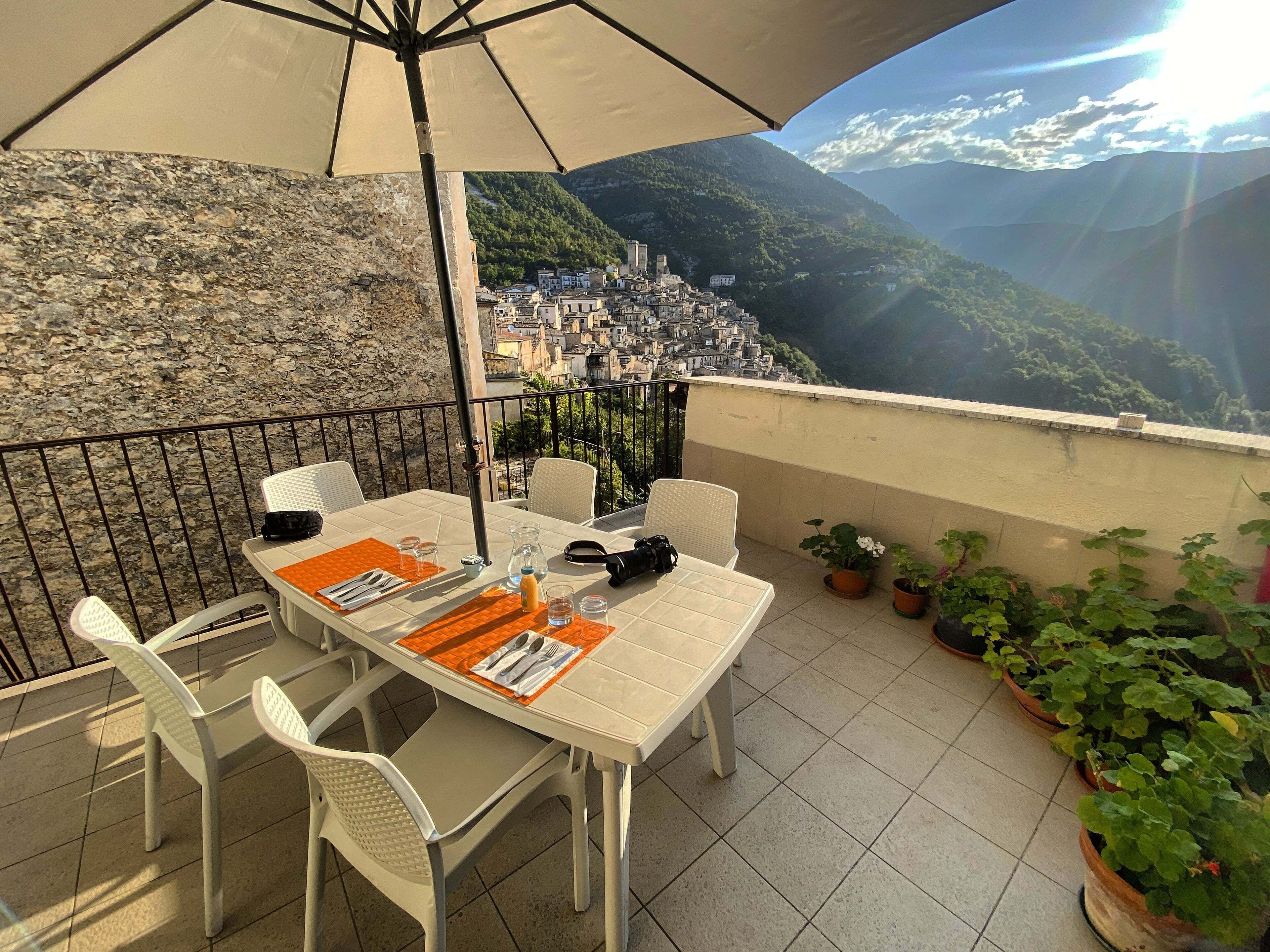 Une terrasse avec une table dressée pour deux personnes, une chaise, un pot de fleurs et une vue sur la ville et les montagnes.