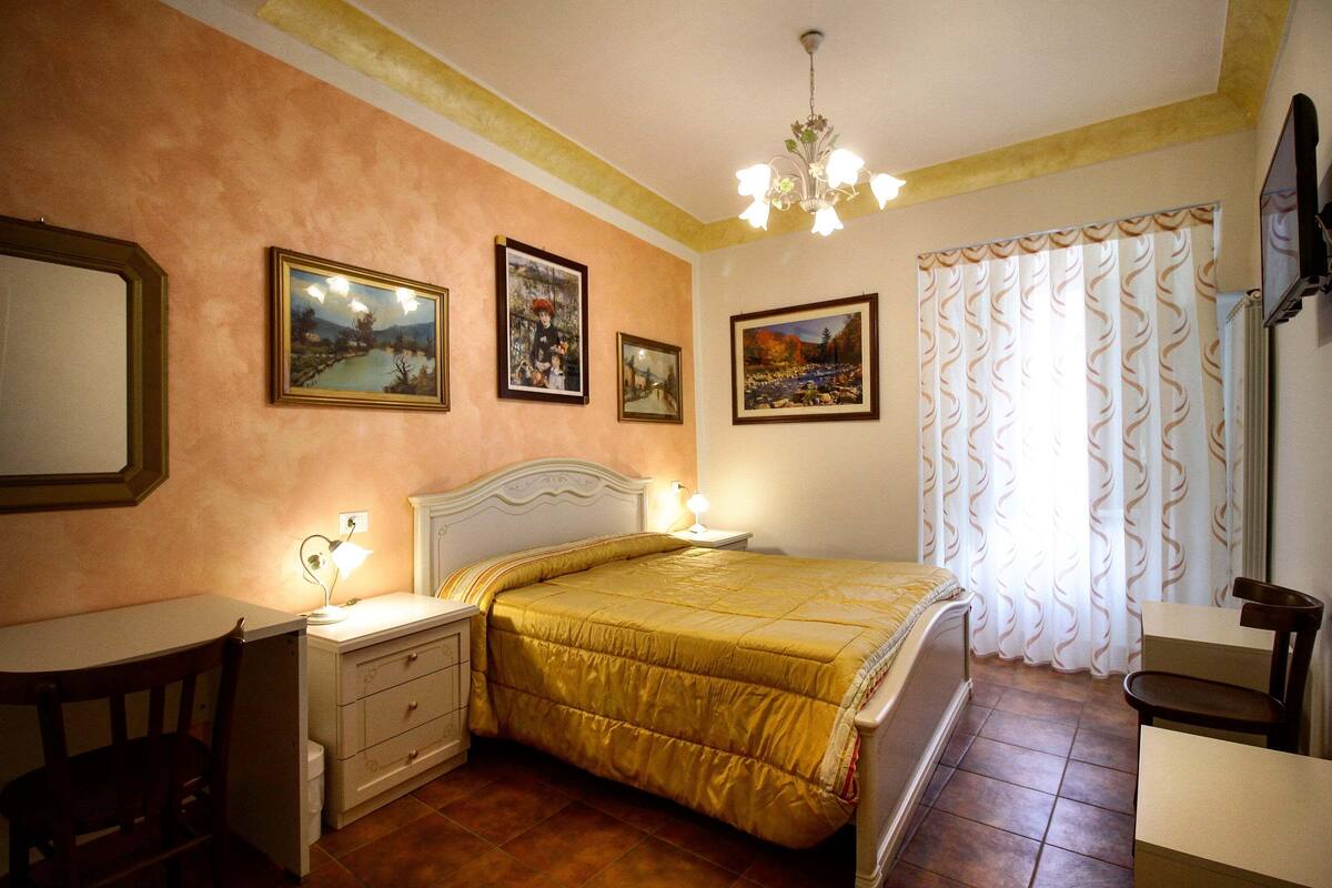 A bedroom with a bed, bedside tables, a desk, and a chair. There are framed pictures on the wall and a window with curtains.