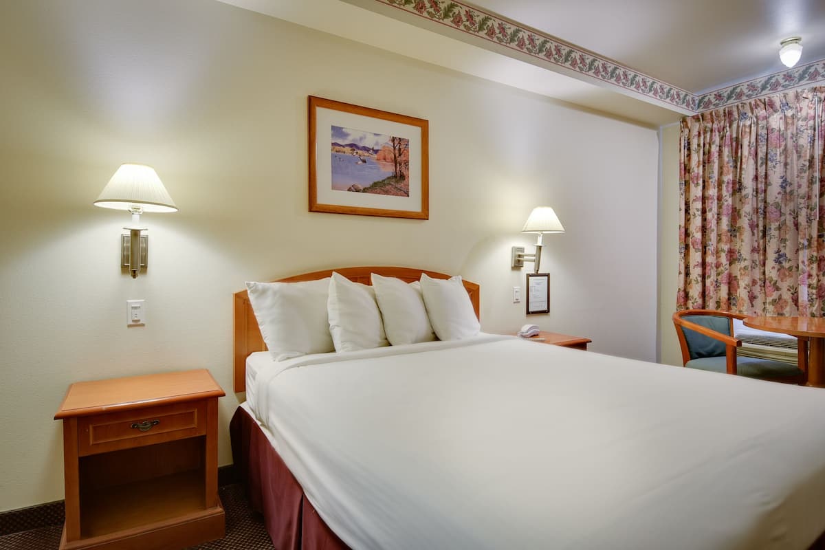 Ein Hotelzimmer mit einem großen Bett, zwei Nachttischlampen, einem kleinen Tisch mit Stuhl und einem blumigen Vorhang.