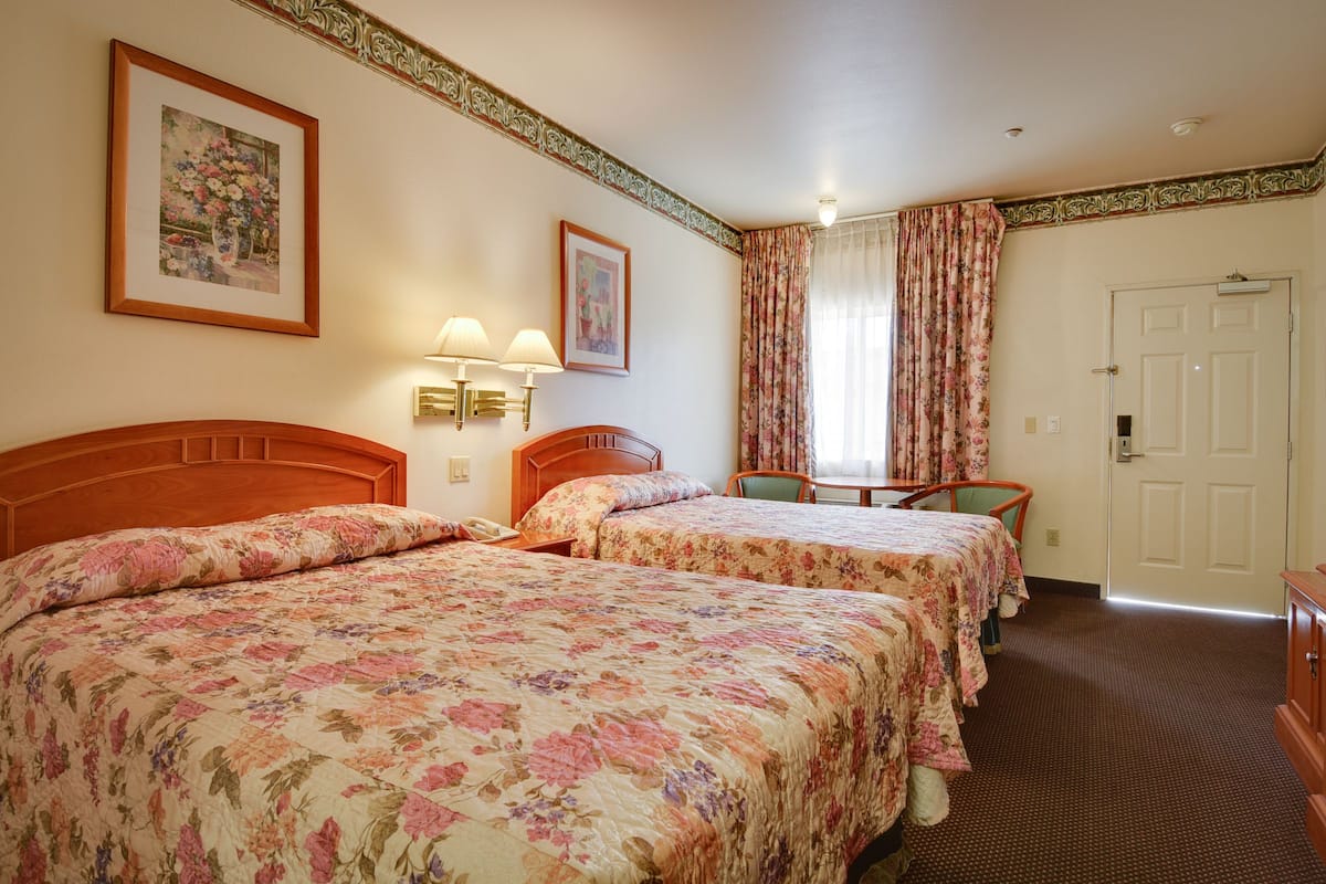 Ein Hotelzimmer mit zwei Betten, einem Schreibtisch und einem Stuhl. Zwei Wandlampen und eine Tür sind vorhanden.