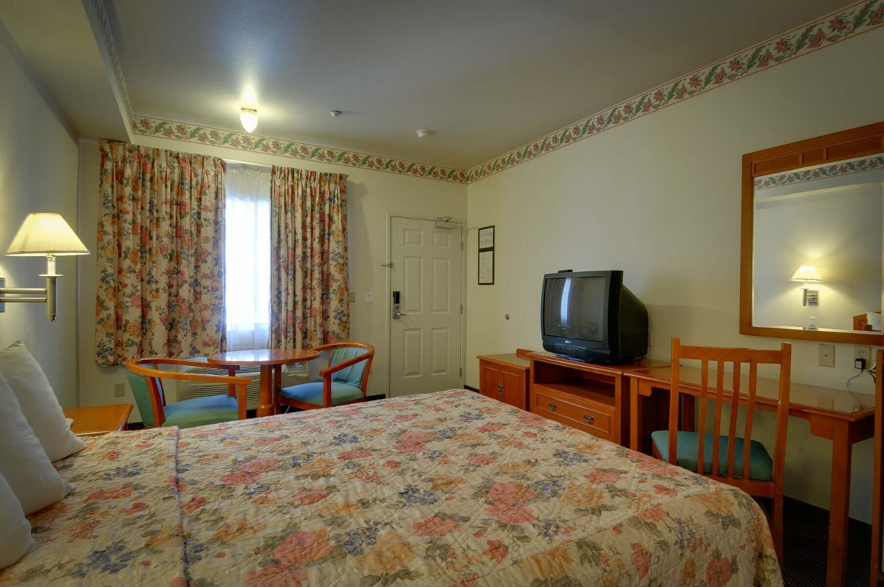A hotel room with a bed, a desk with a television, a chair, and a lamp.