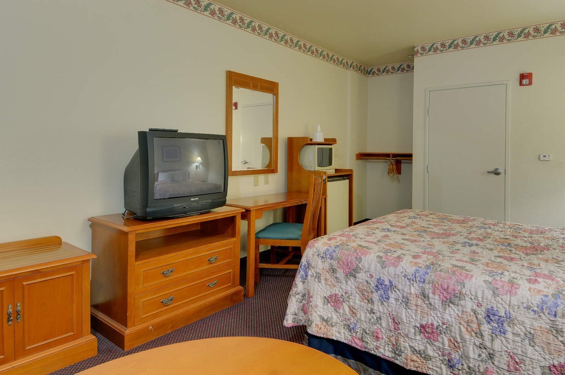 Una habitación de hotel con una cama, un escritorio de madera con televisor, una silla, un aparador y una puerta.