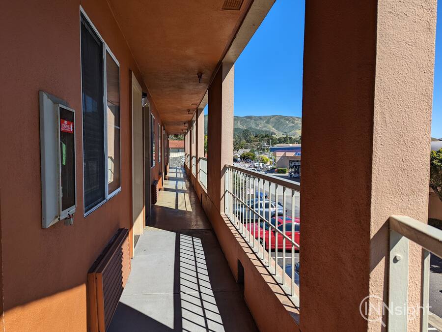 Un long balcon étroit, doté d’une rambarde en métal, qui offre une vue sur un parking et sur des collines lointaines.
