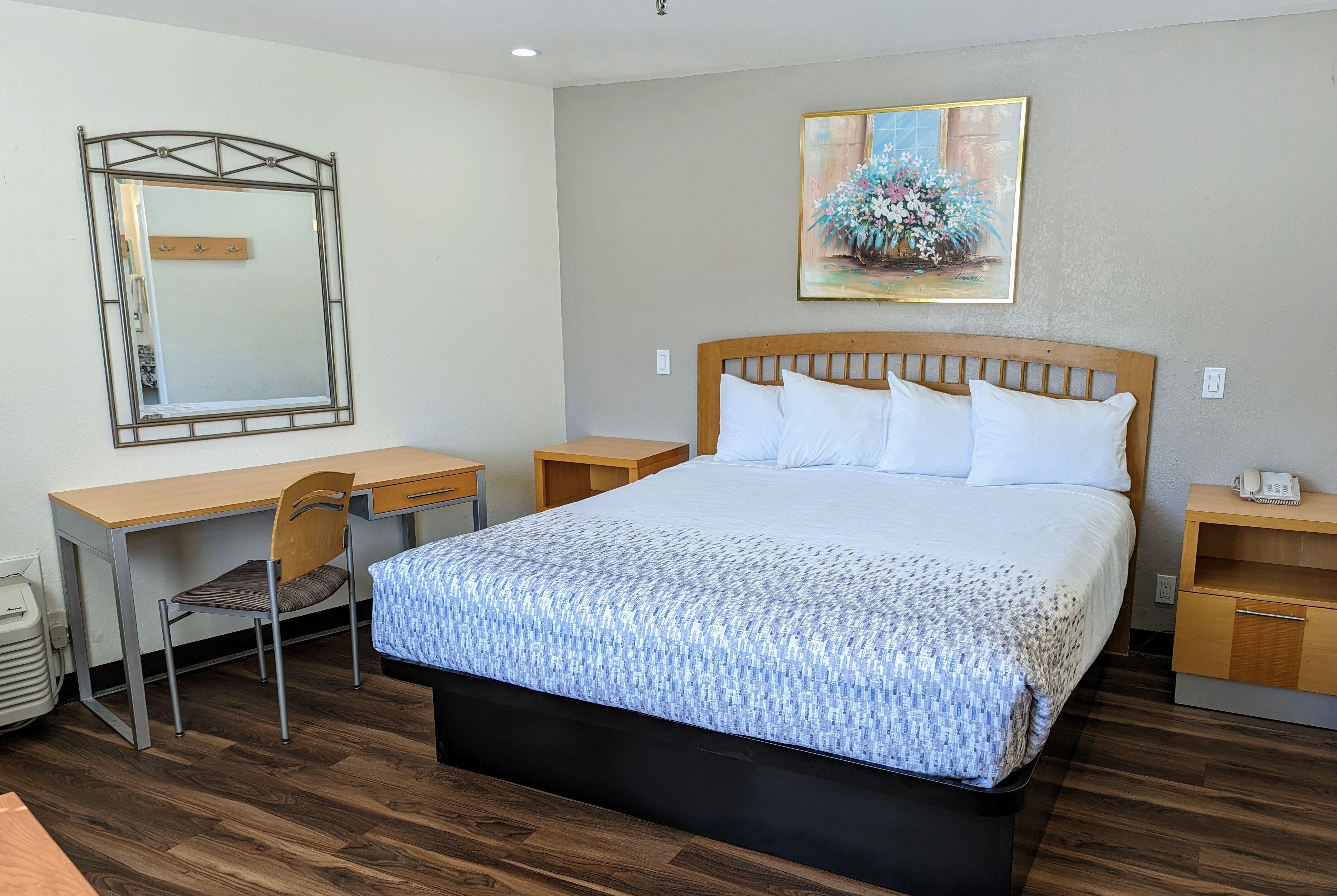 A hotel room with a wooden bed, a desk, a chair, a mirror, and a painting on the wall.