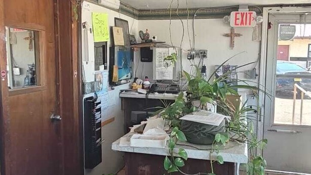 A room with a wooden door, a small desk with a computer, a plant, and a bucket labeled 'Wooly Fresh.'