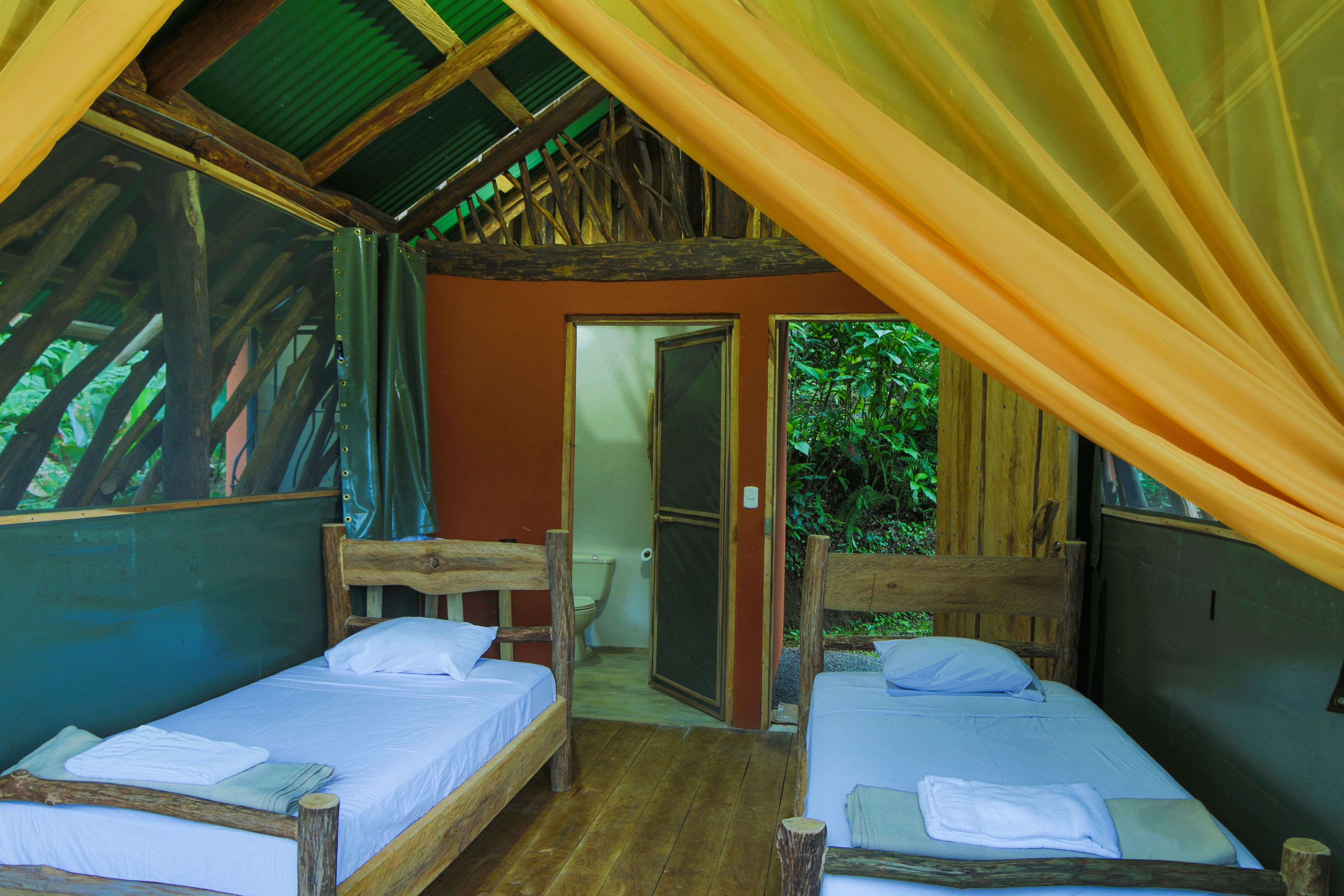 A rustic cabin with two wooden beds, a wooden ceiling, and a door leading to a bathroom.