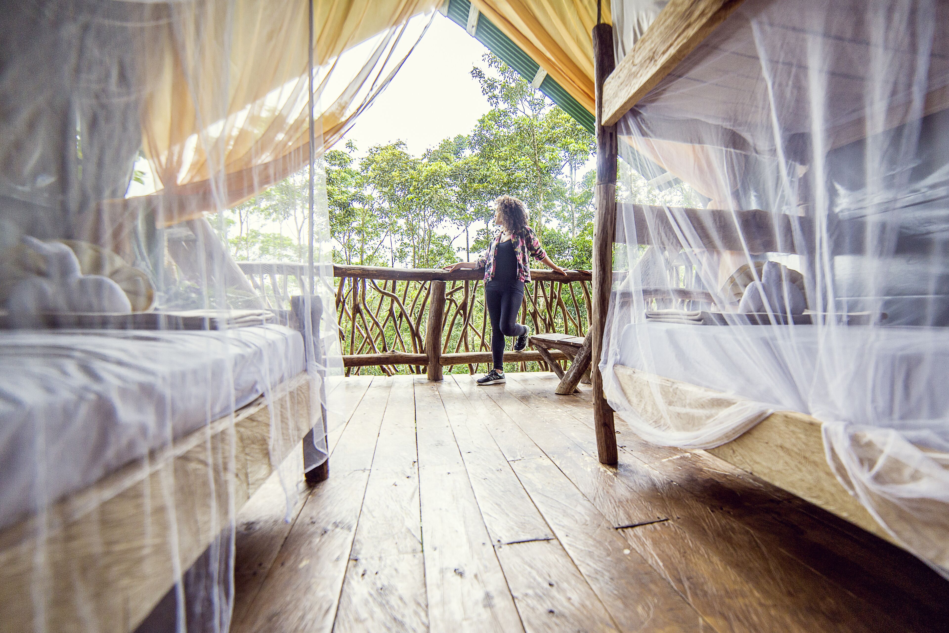 A wooden deck with four beds covered in mosquito netting, a wooden railing, and a view of trees.