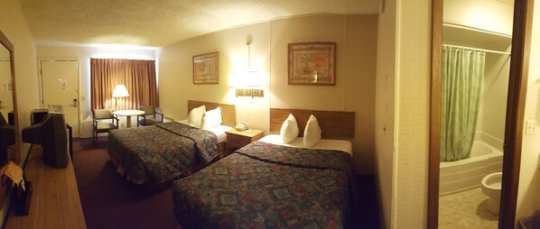 A hotel room with two beds, a desk, a chair, a lamp, and a bathroom with a shower curtain.