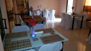 Comedor con mesas dispuestas para cuatro, cubiertas con manteles y posavasos blancos. Hay jarrones con flores sobre las mesas y una lámpara de mesa con luz azul. El cuarto tiene piso de baldosas y una puerta al fondo.
