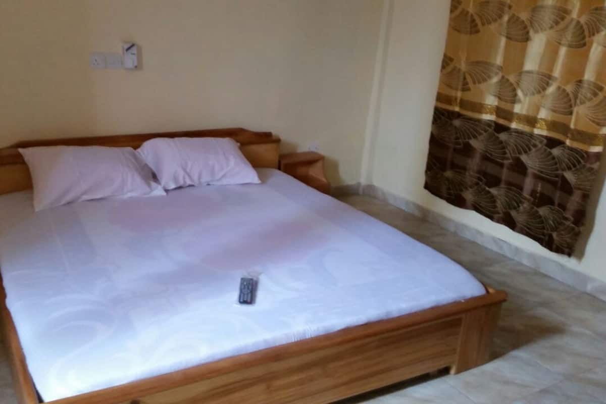 A neatly made bed with a remote control on the mattress, a wooden headboard, and a curtain with a leaf pattern on the window.