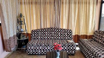 A living room with a patterned sofa, a decorative shelf with figurines, and a curtain backdrop.