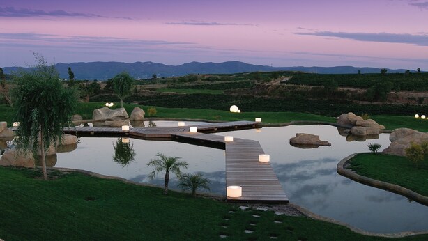 A wooden bridge over a pond with lights, surrounded by greenery and mountains in the background. - Can Bonastre Wine Resort, The Originals Relais (Relais du Silence) (Masquefa)