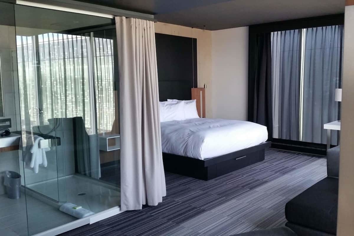 A modern hotel room with a glass shower enclosure, a bed with a dark headboard, a bedside table, and a view of the city through large windows.