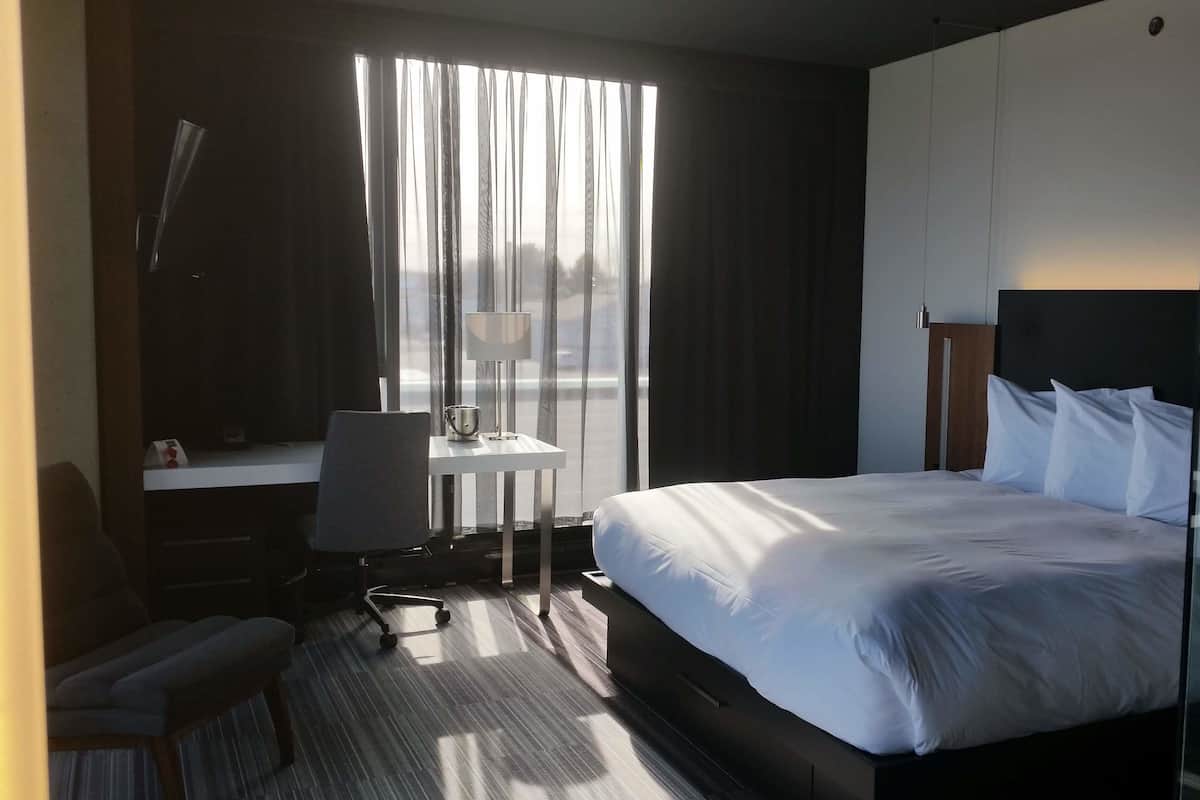 A hotel room with a large bed, a desk with a chair, a small table with a lamp, and a view through the window.